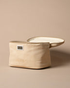 A cream coloured canvas milk pouch with a small digital temperature indicator window and embossed brand name, shown next to its matching lid on a solid beige background.