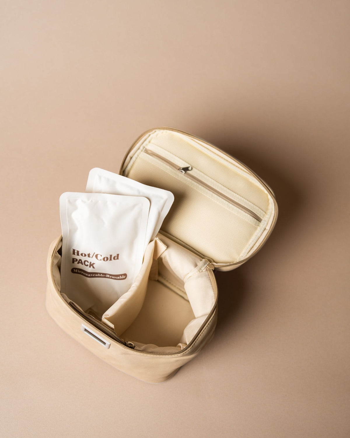 An open beige zippered pouch or organizer bag on a warm beige background, with two white sachets inside. One sachet has visible text reading 'Hot/cold PACK' with smaller text below.
