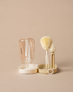 A baby bottle cleaning set on a solid beige background, featuring a clear bottle, a beige brush with a white bristle head, a small scrubber, and a small brush in a matching beige and white holder.
