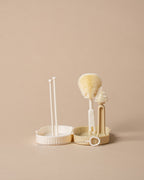 A set of three cream-colored plastic cleaning tools, including a small brush, a scrubber, and a stand with two applicator sticks, arranged on a solid beige background.