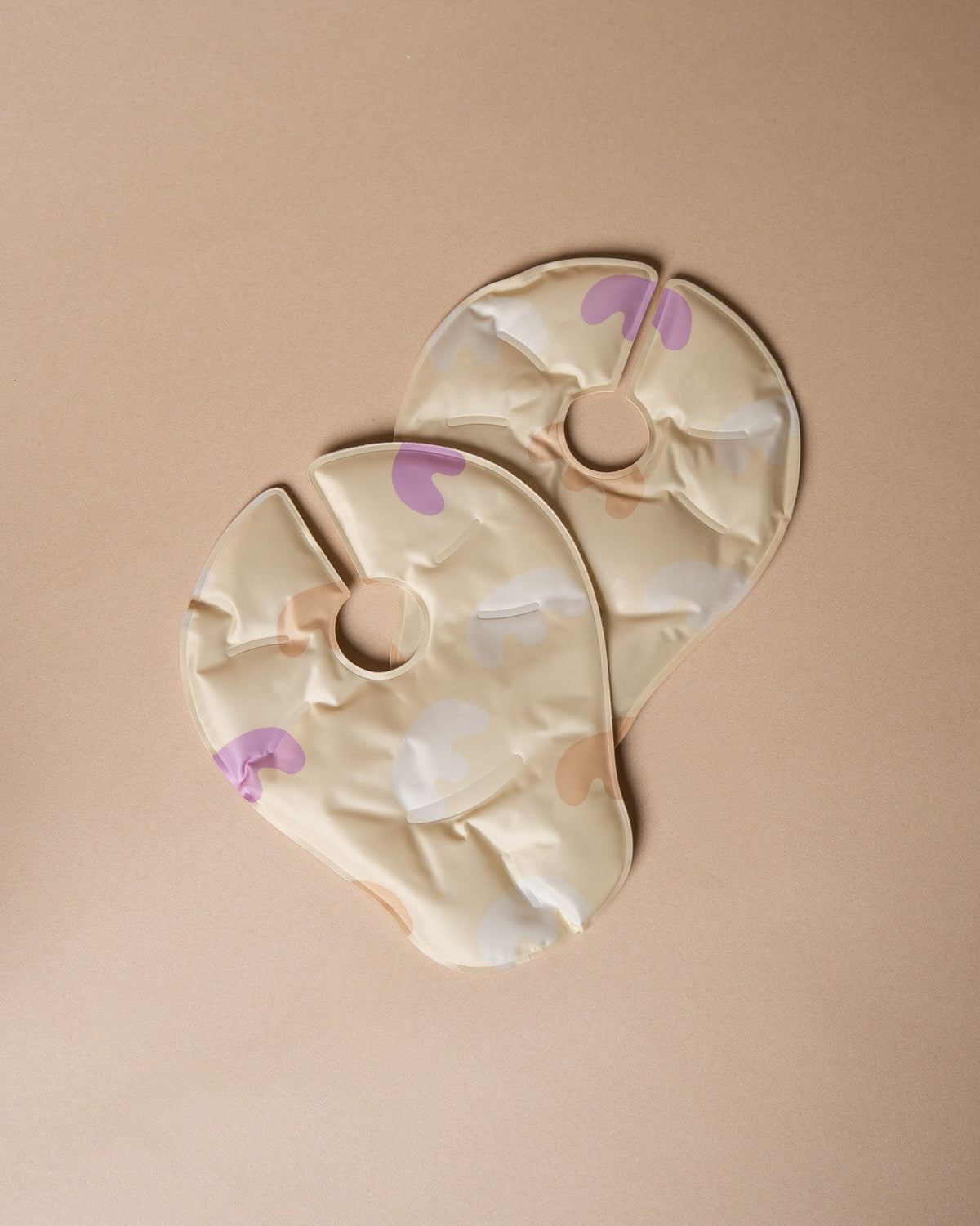 Two beige baby nursing pads with a purple and white abstract pattern, arranged on a solid beige background.